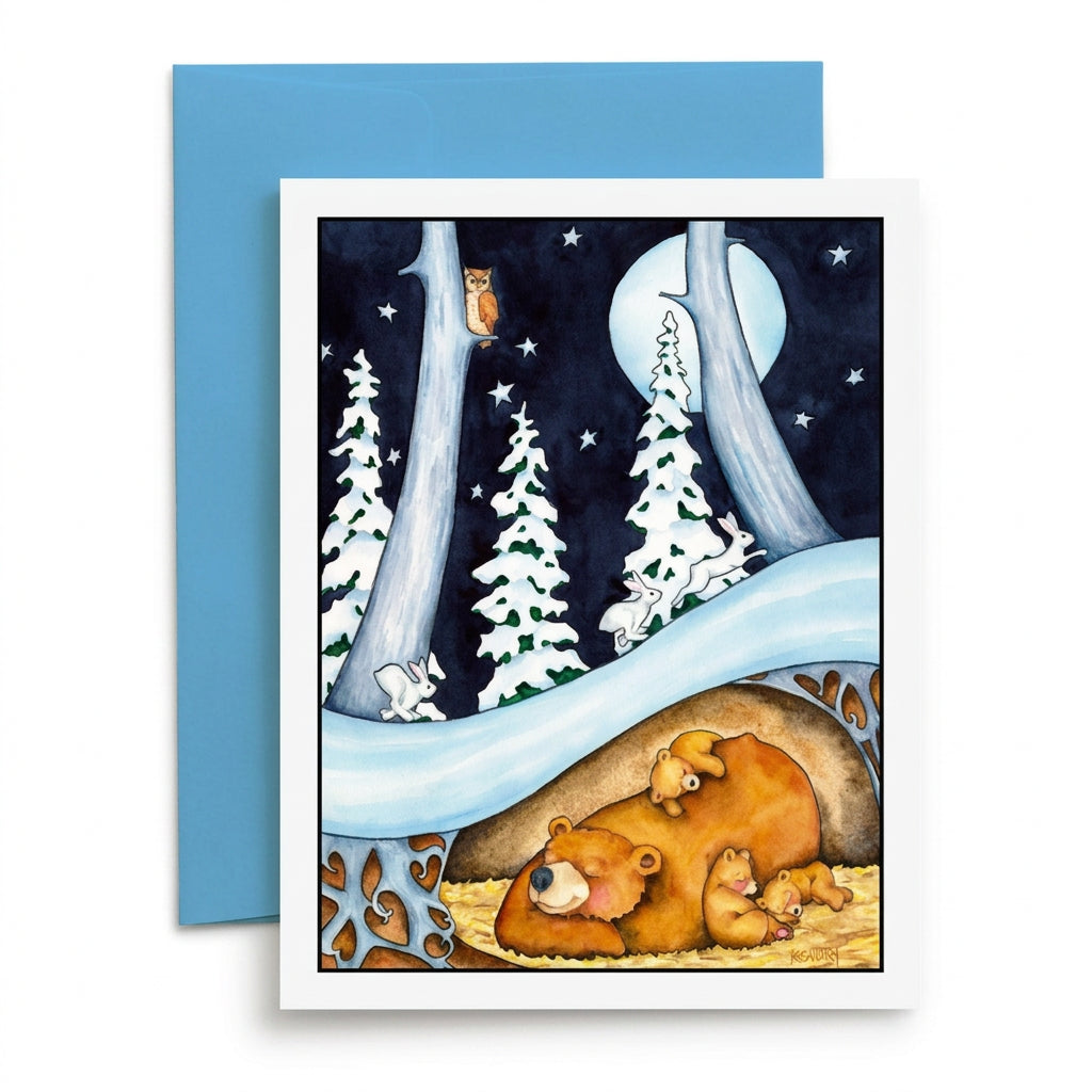 A greeting card with a whimsical forest illustration, shown with a blue envelope on a light wood surface. The card features a sleeping bear family under a tree, a slide, snowy trees, and a owl in a moonlit night scene.