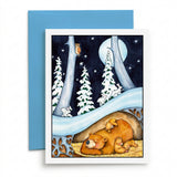 A greeting card with a whimsical forest illustration, shown with a blue envelope on a light wood surface. The card features a sleeping bear family under a tree, a slide, snowy trees, and a owl in a moonlit night scene.