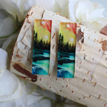 Pair of earrings with nature-themed design on a wooden block with flowers in the background