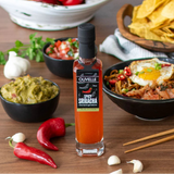 Olivelle Spicy Sriracha bottle on a wooden table with food items like guacamole, chips, and noodles.
