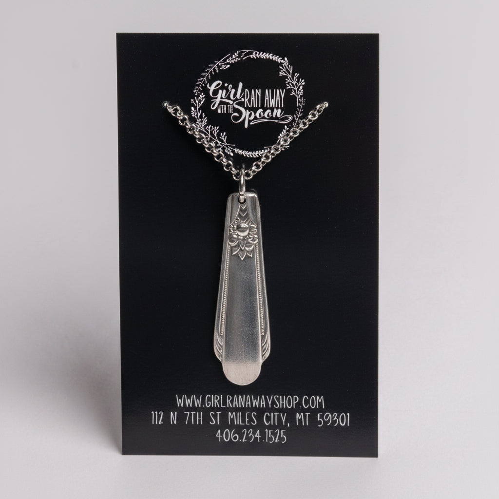 Silver pendant necklace on a black card with branding and contact information.