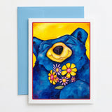 A square art print or card featuring a colorful watercolor-style bear illustration, placed on top of a blue envelope against a light wood surface. The bear is predominantly blue with a yellow face and yellow flowers in front, and the print has a white border with a thin red inner border.