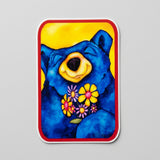 A rectangular art print or sticker with rounded corners featuring a colorful illustration of a blue bear holding yellow and pink flowers, set against a yellow background and bordered in red, shown on a light gray textured surface.