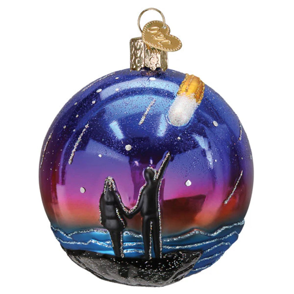 Decorative glass ornament with two figures on a rock, one holding a candle, against a colorful sky.