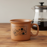 Brown mug with mountain design on a wooden table with a blurred coffee press in the background