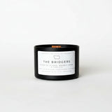 A black glass jar candle with a white label reading 'THE BRIDGERS' and a small copper-colored wick, set on a white background.