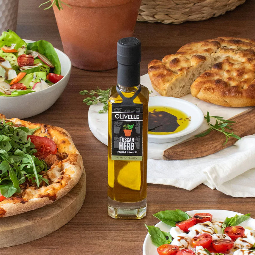 Olivelle Tuscan Herb olive oil bottle on a wooden table with pizza, salad, and bread.