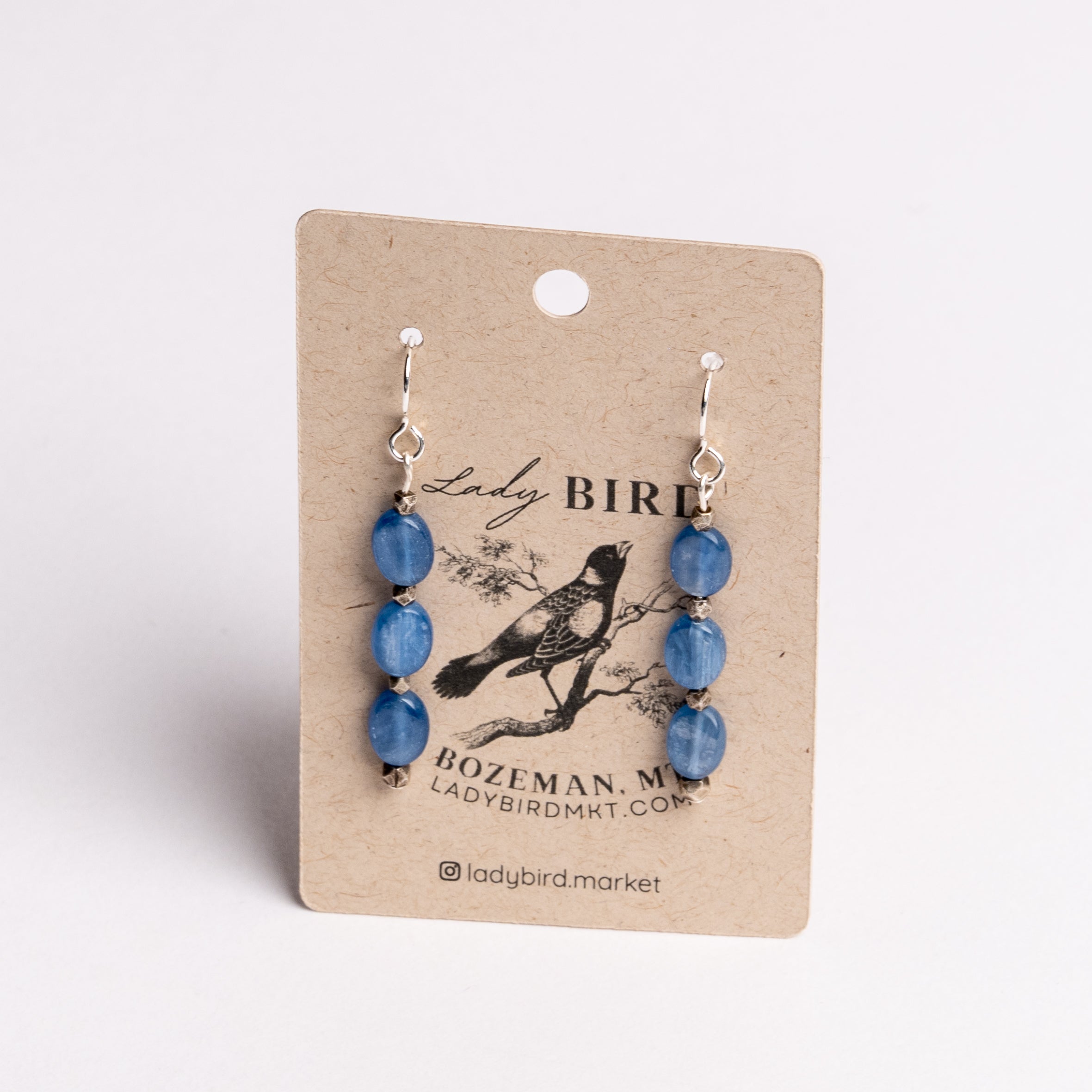A pair of dangling earrings with blue faceted beads and silver hooks, displayed on a kraft paper card printed with the brand name Lady Bird.