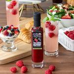 Olivelle raspberry vinegar bottle with glasses of raspberry-laced drinks and snacks on a wooden table.