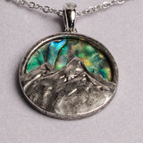 A silver oval pendant necklace featuring a textured mountain design over a vibrant green and blue abalone shell inlay, shown on a light gray background.