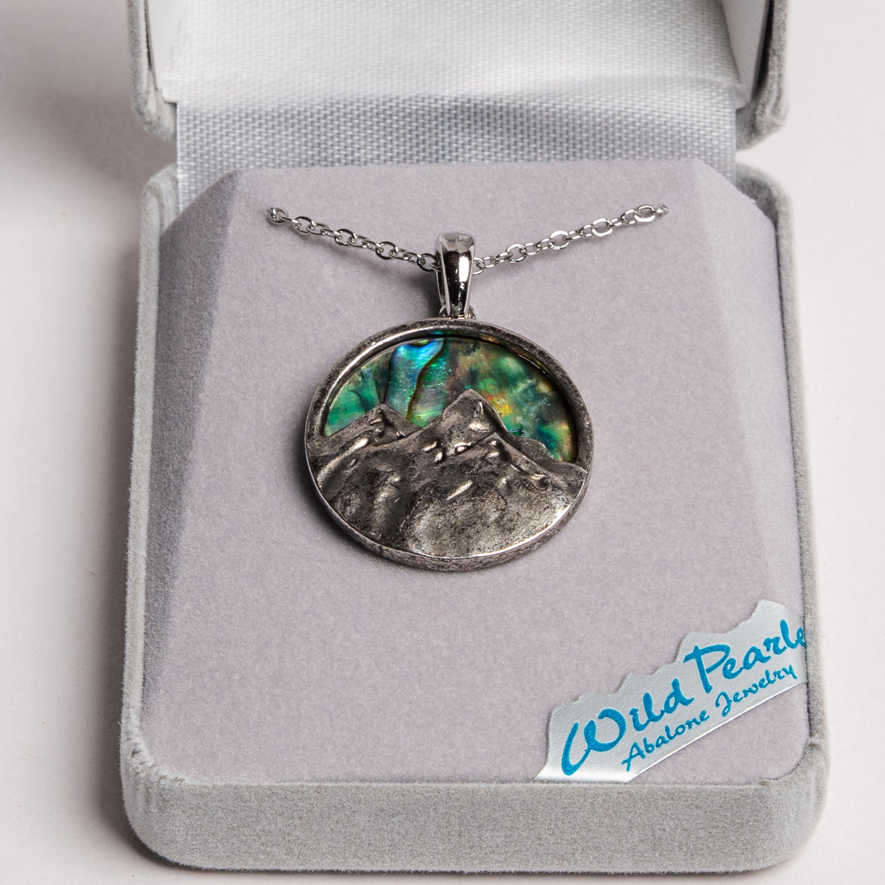A silver pendant necklace featuring a mountain landscape design over an iridescent abalone shell inlay, displayed inside a gray jewelry box. A 'Wild Pearls Abalone Jewelry' brand tag is visible on the box.