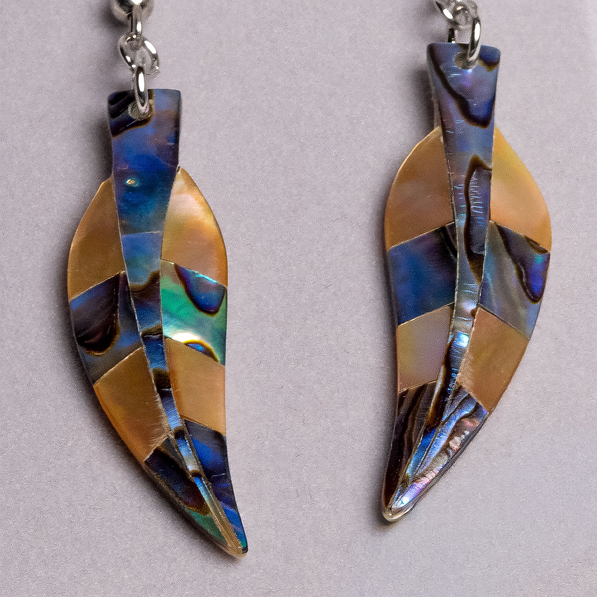 A pair of leaf-shaped dangle earrings made from iridescent abalone shell pieces in blue, gold, and brown tones, hanging from silver-toned rings on a light gray background.