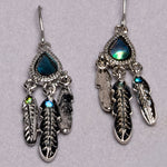 A pair of silver-toned dangle earrings with teardrop-shaped iridescent blue-green stones and three feather charms, including one larger feather with a small green stone, displayed on a light gray textured background.