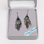 A pair of silver-toned dreamcatcher-style earrings with iridescent blue-green teardrop stones and feather charms, displayed in an open gray jewelry box. A small card reading 'Wild Pearls Hypo Allergenic Posts' is visible inside the box.