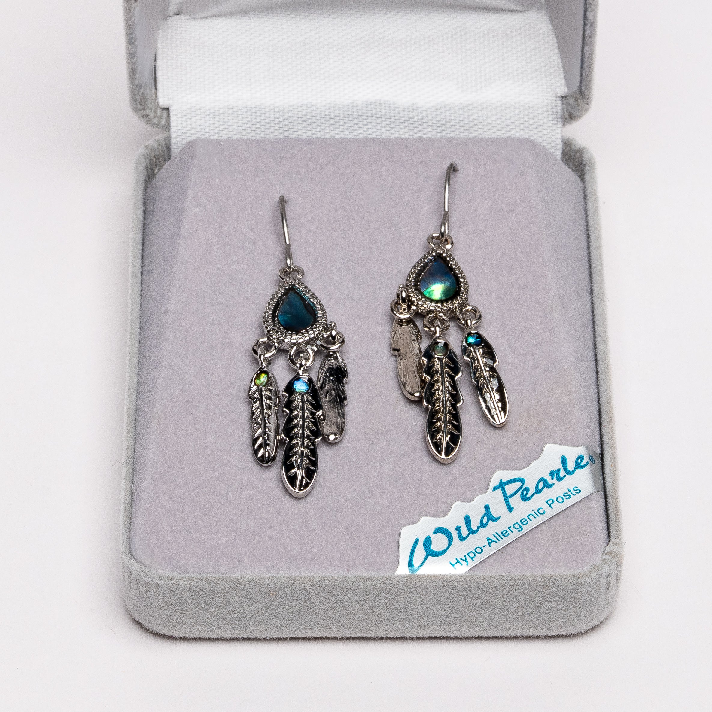 A pair of silver-toned dreamcatcher-style earrings with iridescent blue-green teardrop stones and feather charms, displayed in an open gray jewelry box. A small card reading 'Wild Pearls Hypo Allergenic Posts' is visible inside the box.