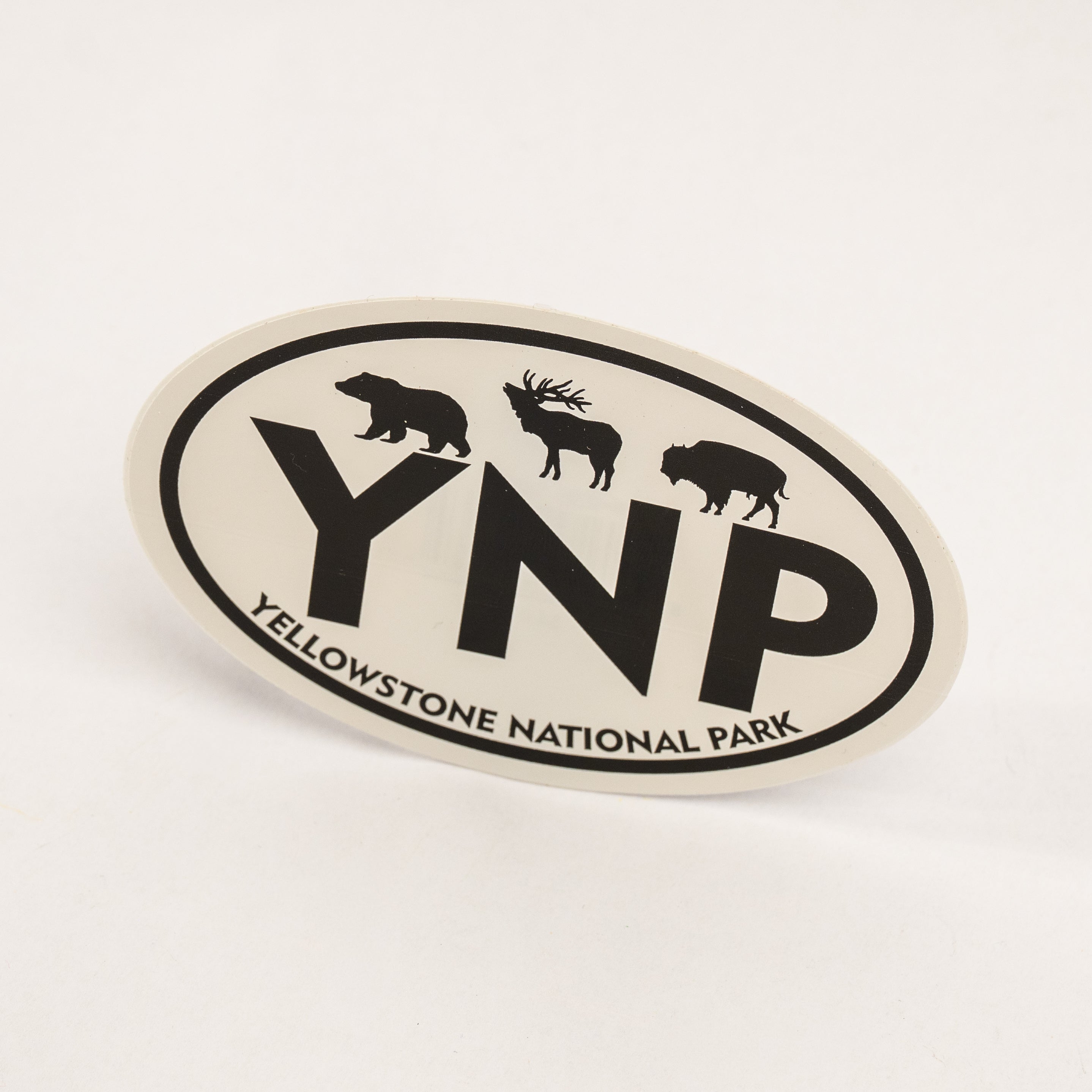 An oval Yellowstone National Park sticker featuring black silhouettes of a bear, moose, and bison above the large letters 'YNP', with 'YELLOWSTONE NATIONAL PARK' curved along the bottom border, shown on a clean white background.