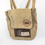 A beige canvas tote bag with dark brown accents, featuring a front pocket with a brown patch that reads 'America's First National Park' and a circular Yellowstone logo, alongside vertical 'Yellowstone' text.