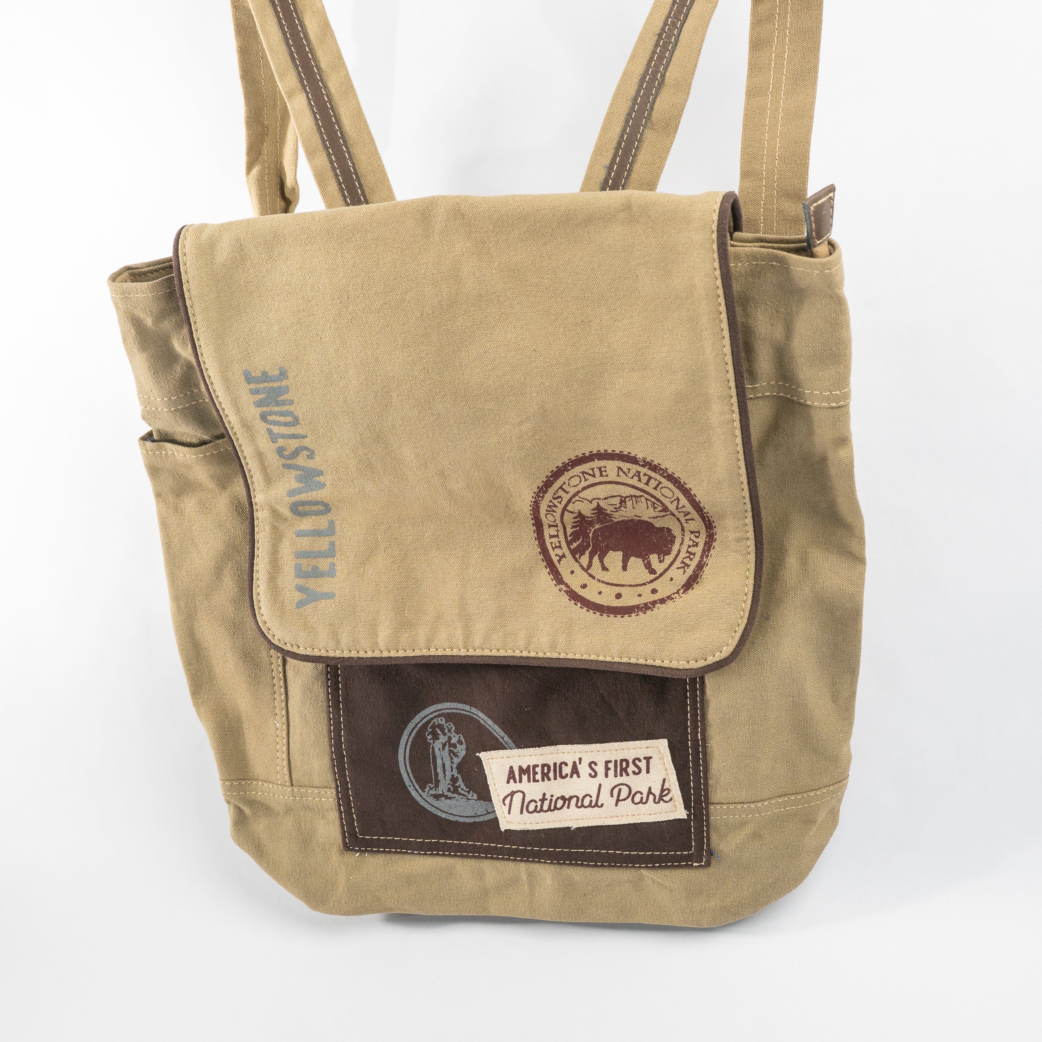 A beige canvas tote bag with dark brown accents, featuring a front pocket with a brown patch that reads 'America's First National Park' and a circular Yellowstone logo, alongside vertical 'Yellowstone' text.