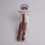 A packaged bag of rock candy on a plain light background. The clear plastic bag contains three long wooden sticks coated with dark purple crystalline candy, and the top card reads 'Old Fashioned Wild Huckleberry Rock Candy' with purple mountain 