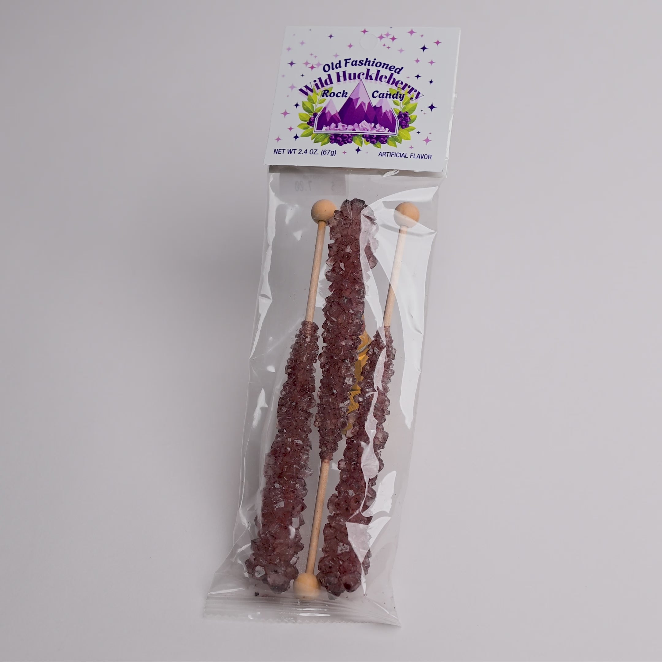 A packaged bag of rock candy on a plain light background. The clear plastic bag contains three long wooden sticks coated with dark purple crystalline candy, and the top card reads 'Old Fashioned Wild Huckleberry Rock Candy' with purple mountain 