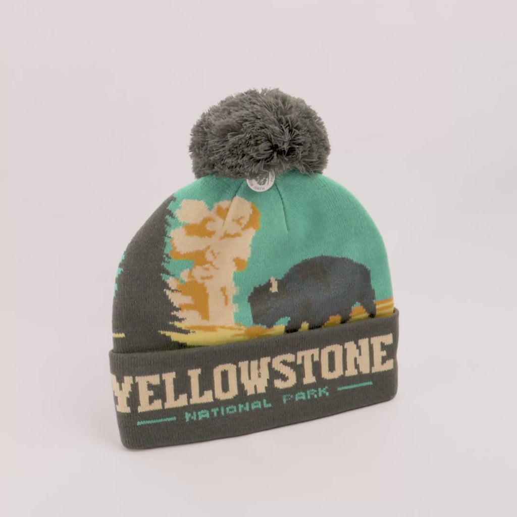 A knit Yellowstone National Park pom-pom beanie featuring a turquoise and olive green design with a bison and mushroom cloud graphic, and a folded cuff reading 'YELLOWSTONE NATIONAL PARK'.