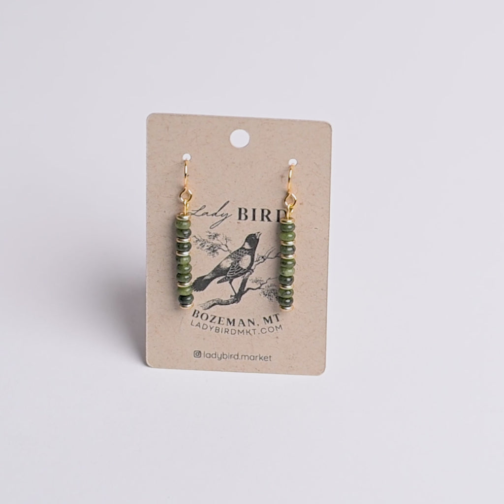 A pair of dangling earrings with green faceted beads and gold hooks, displayed on a kraft paper card printed with the brand name Lady Bird.