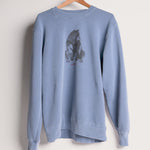 A dusty blue crewneck sweatshirt hanging on a wooden hanger against a white wall. The front of the sweatshirt features a dark gray watercolor-style graphic of a bear sitting on its hind legs, with small red text underneath