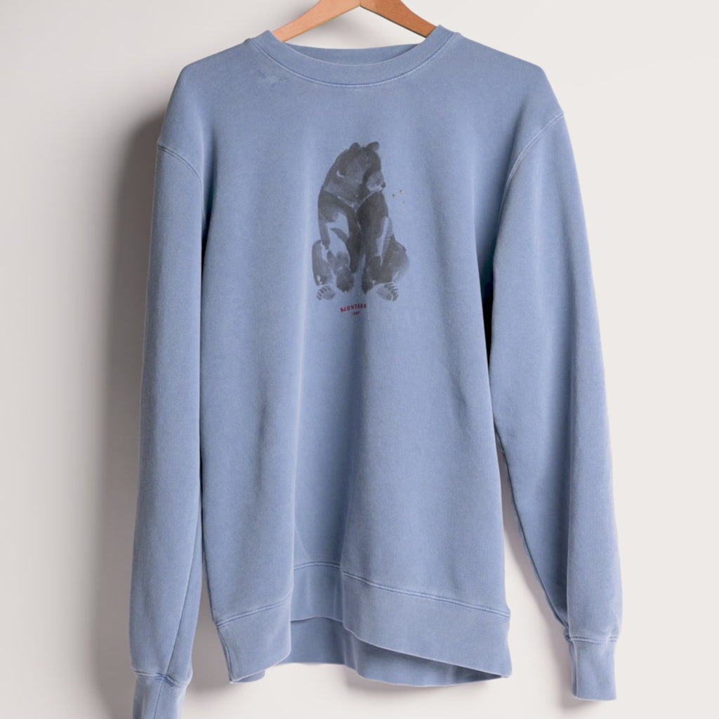 A dusty blue crewneck sweatshirt hanging on a wooden hanger against a white wall. The front of the sweatshirt features a dark gray watercolor-style graphic of a bear sitting on its hind legs, with small red text underneath