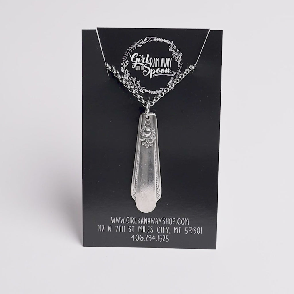 Silver pendant necklace on a black card with branding and contact information.