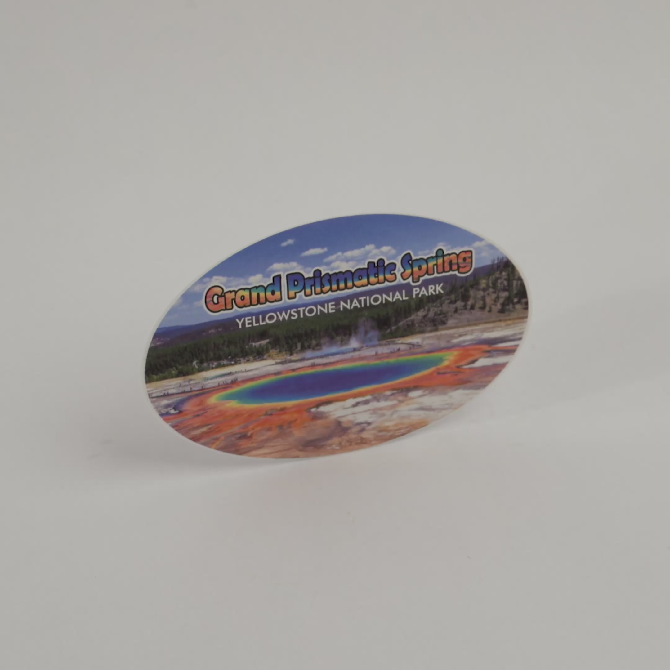 An oval-shaped souvenir sticker featuring a colorful landscape of Grand Prismatic Spring in Yellowstone National Park, displayed on a plain white background.