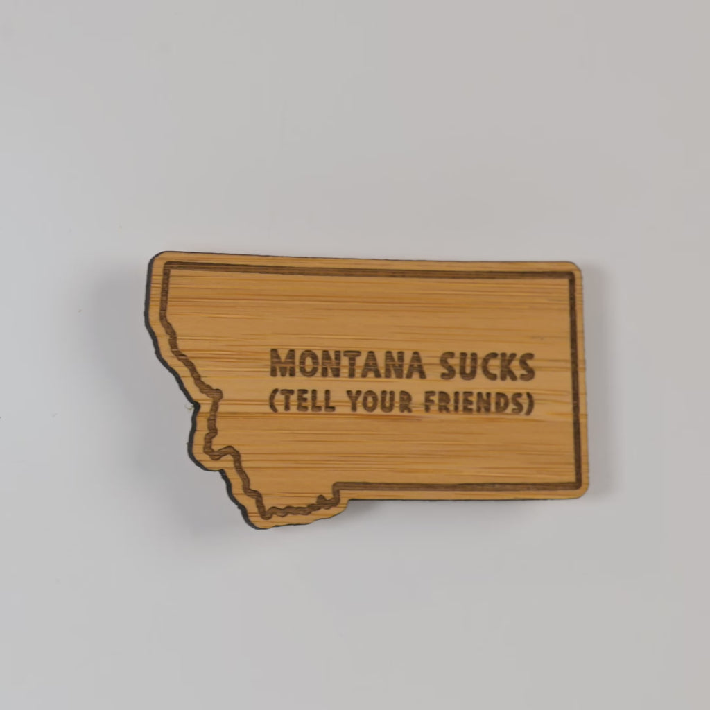 Wooden state-shaped sign with 'Montana Sucks (Tell Your Friends)' text on a white background