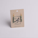 A pair of dangling earrings with green and gold spacer beads and gold hooks, displayed on a brown kraft paper card with 'Lady Bird' branding.