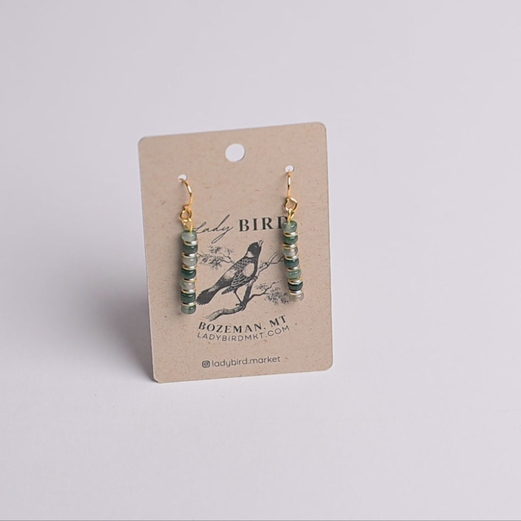 A pair of dangling earrings with green and gold spacer beads and gold hooks, displayed on a brown kraft paper card with 'Lady Bird' branding.