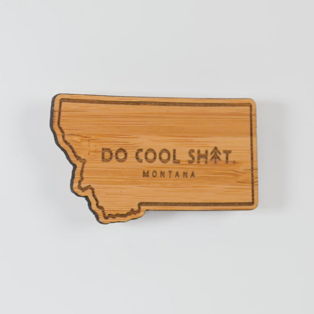 Wooden state-shaped magnet with 'Do Cool Sh*t. Montana' text on a white background