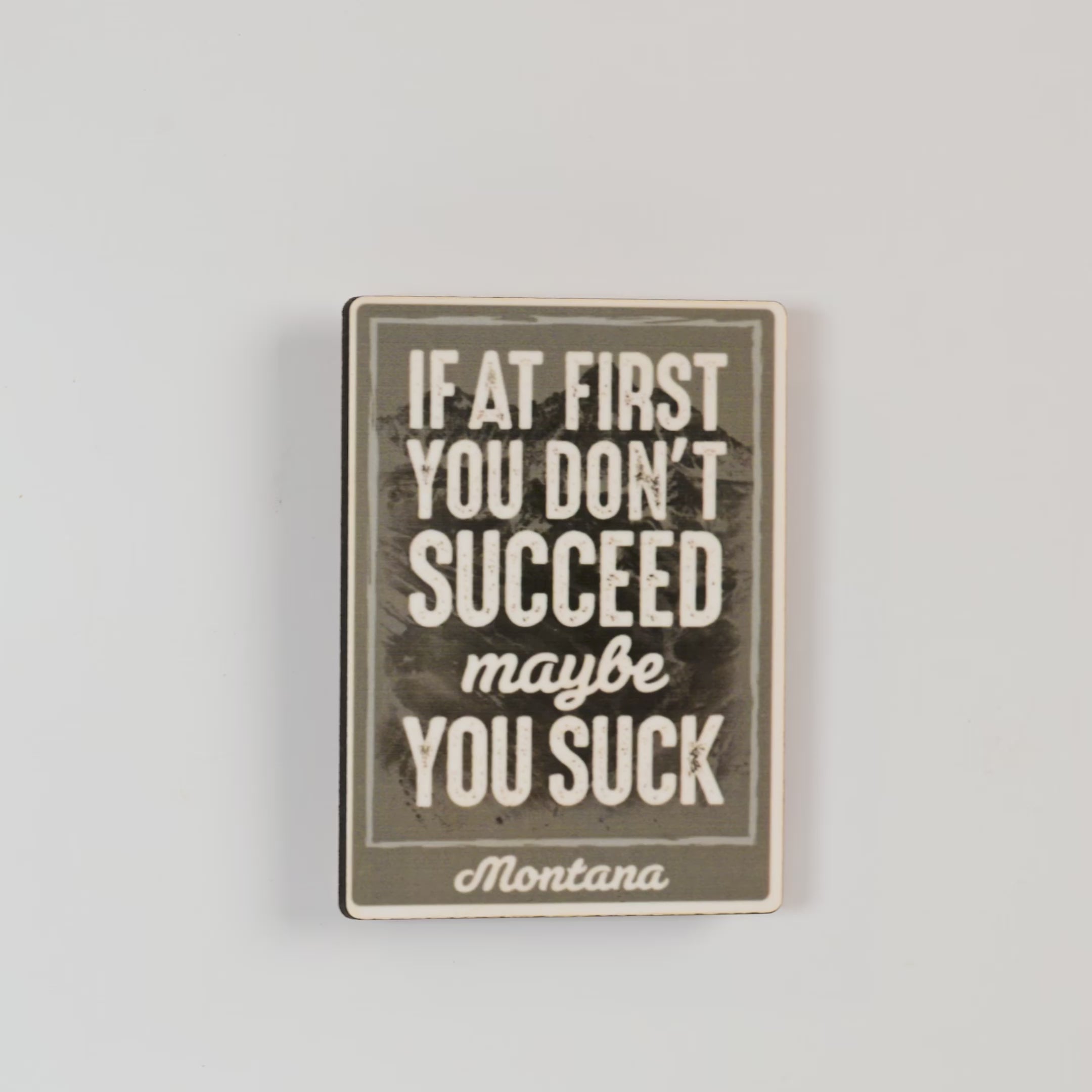 Motivational magnet with text 'If at first you don't succeed, maybe you suck. Montana' on a textured background.