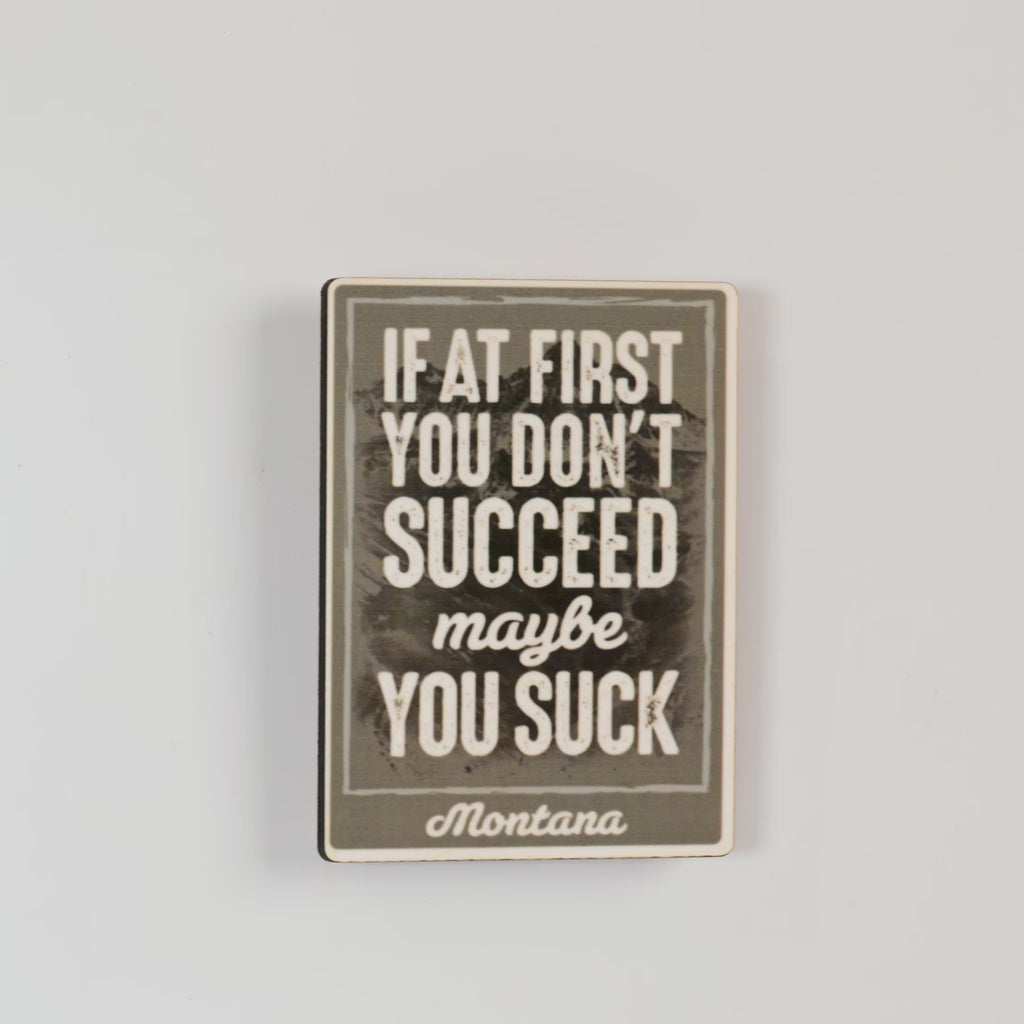 Motivational magnet with text 'If at first you don't succeed, maybe you suck. Montana' on a textured background.