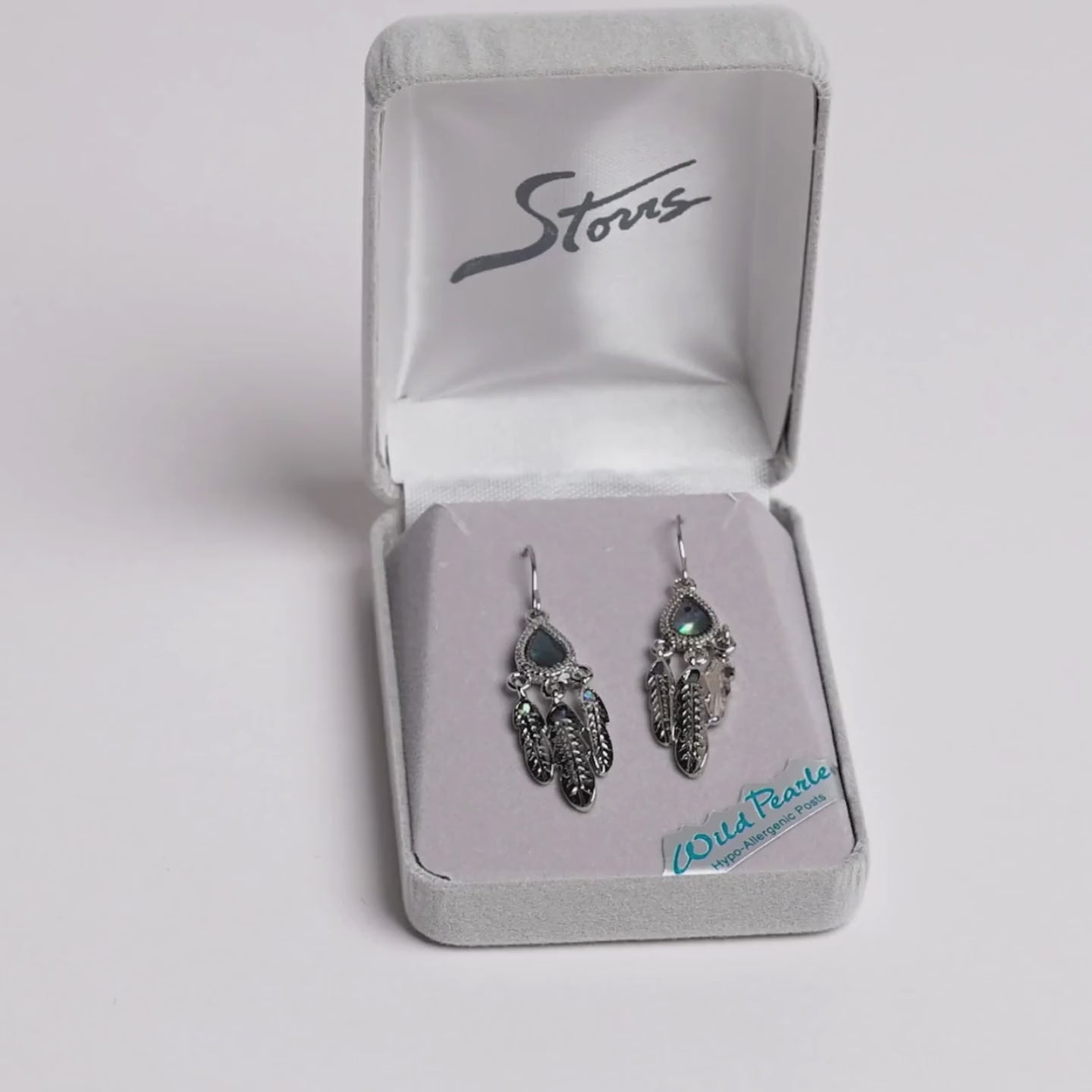 A pair of silver-toned dreamcatcher-style earrings with iridescent blue-green teardrop stones and feather charms, displayed in an open gray jewelry box. A small card reading 'Wild Pearls Hypo Allergenic Posts' is visible inside the box.