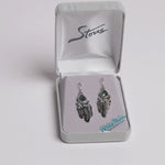 A pair of silver-toned dreamcatcher-style earrings with iridescent blue-green teardrop stones and feather charms, displayed in an open gray jewelry box. A small card reading 'Wild Pearls Hypo Allergenic Posts' is visible inside the box.