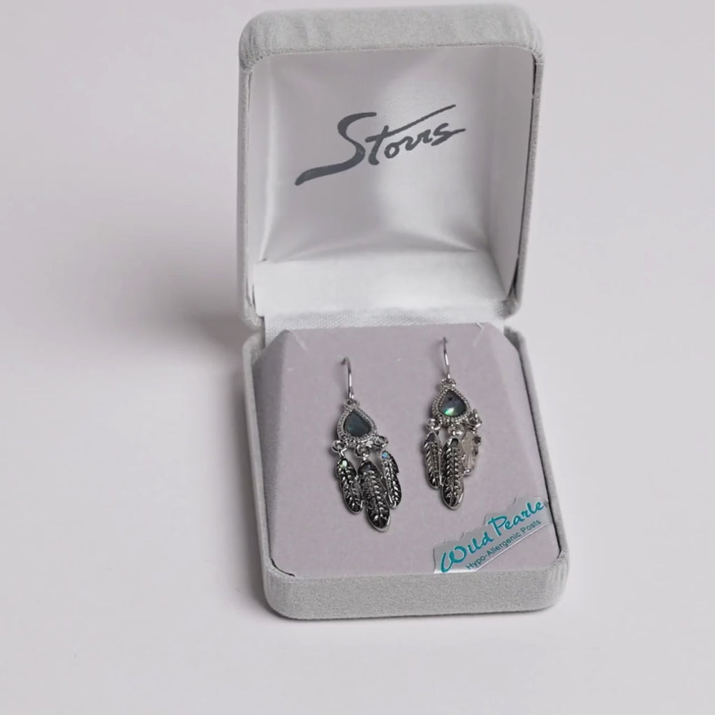 A pair of silver-toned dreamcatcher-style earrings with iridescent blue-green teardrop stones and feather charms, displayed in an open gray jewelry box. A small card reading 'Wild Pearls Hypo Allergenic Posts' is visible inside the box.