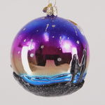 Decorative glass ornament with two figures on a rock, one holding a candle, against a colorful sky.