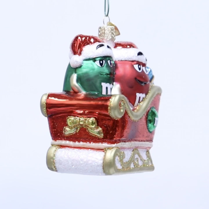 A Christmas ornament featuring two M&M's characters in a red sleigh. The green M&M is on the left wearing a Santa hat, and the red M&M is on the right with a Santa hat.