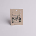 A pair of dangling earrings displayed on a kraft paper card. The earrings have silver fishhook wires and cylindrical beads featuring black, turquoise blue, and white stripes, with a small black bird illustration in the center.