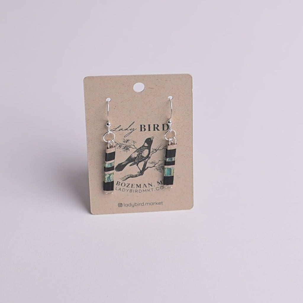 A pair of dangling earrings displayed on a kraft paper card. The earrings have silver fishhook wires and cylindrical beads featuring black, turquoise blue, and white stripes, with a small black bird illustration in the center.