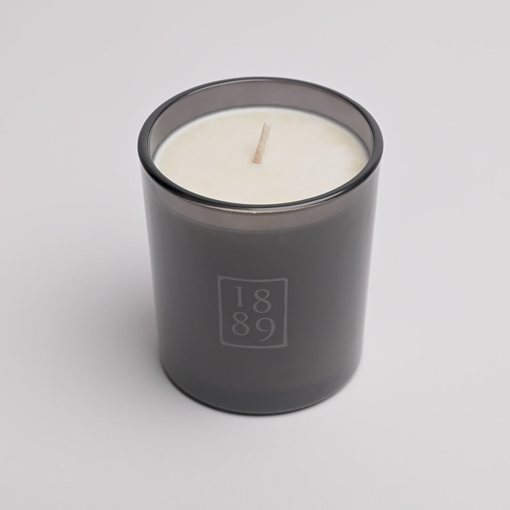 A single-wick scented candle in a dark smoky gray glass jar with a white wax fill and a light wood wick. The front of the jar features a frosted gray square logo containing the numbers '1889'.