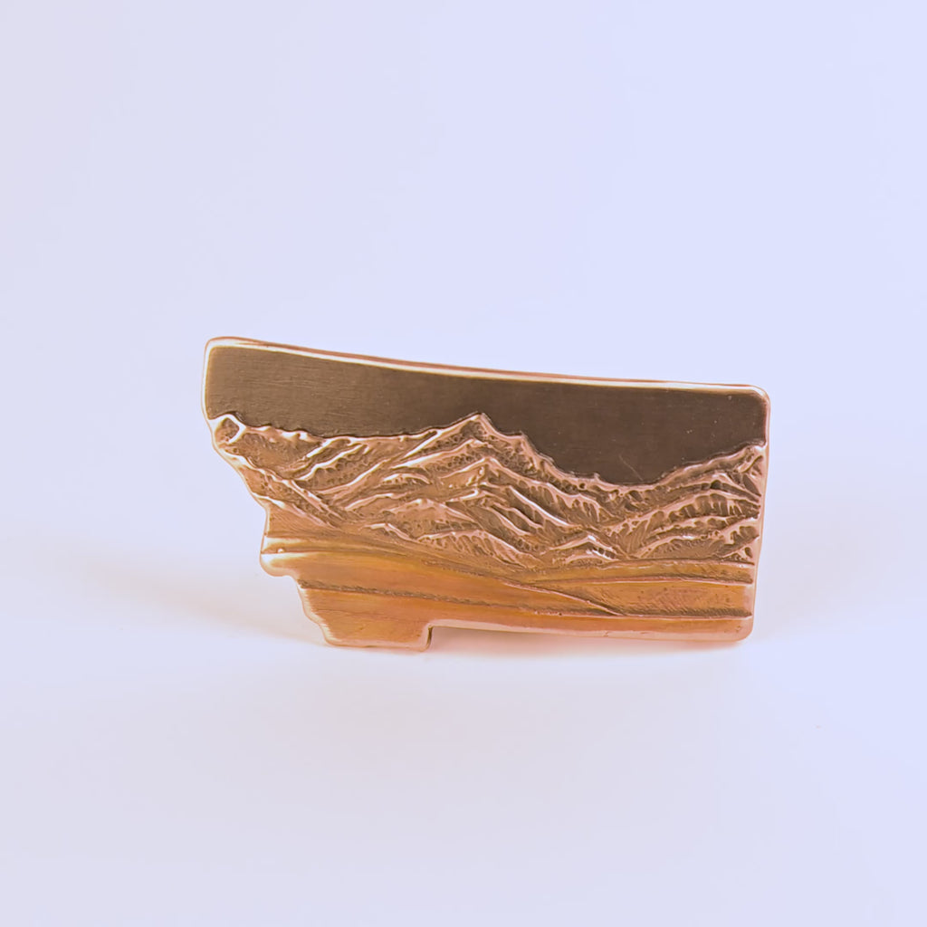 Metallic bronze belt buckle shaped like Montana with mountain design on a white background