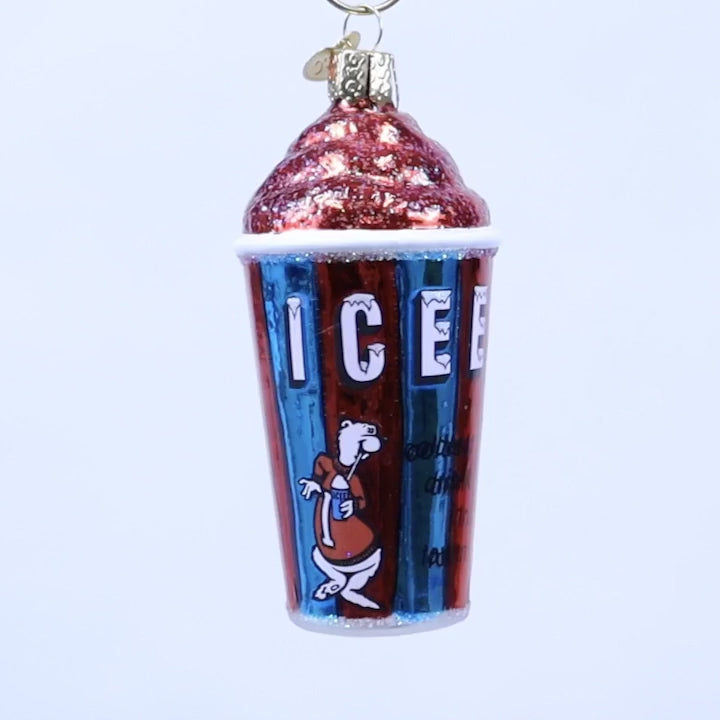 A novelty Christmas ornament shaped like a cup of iced coffee, featuring a red glittery swirl top, a red and blue striped cup with the word 'ICE' and a cartoon character, and a gold hanging loop on a white background.