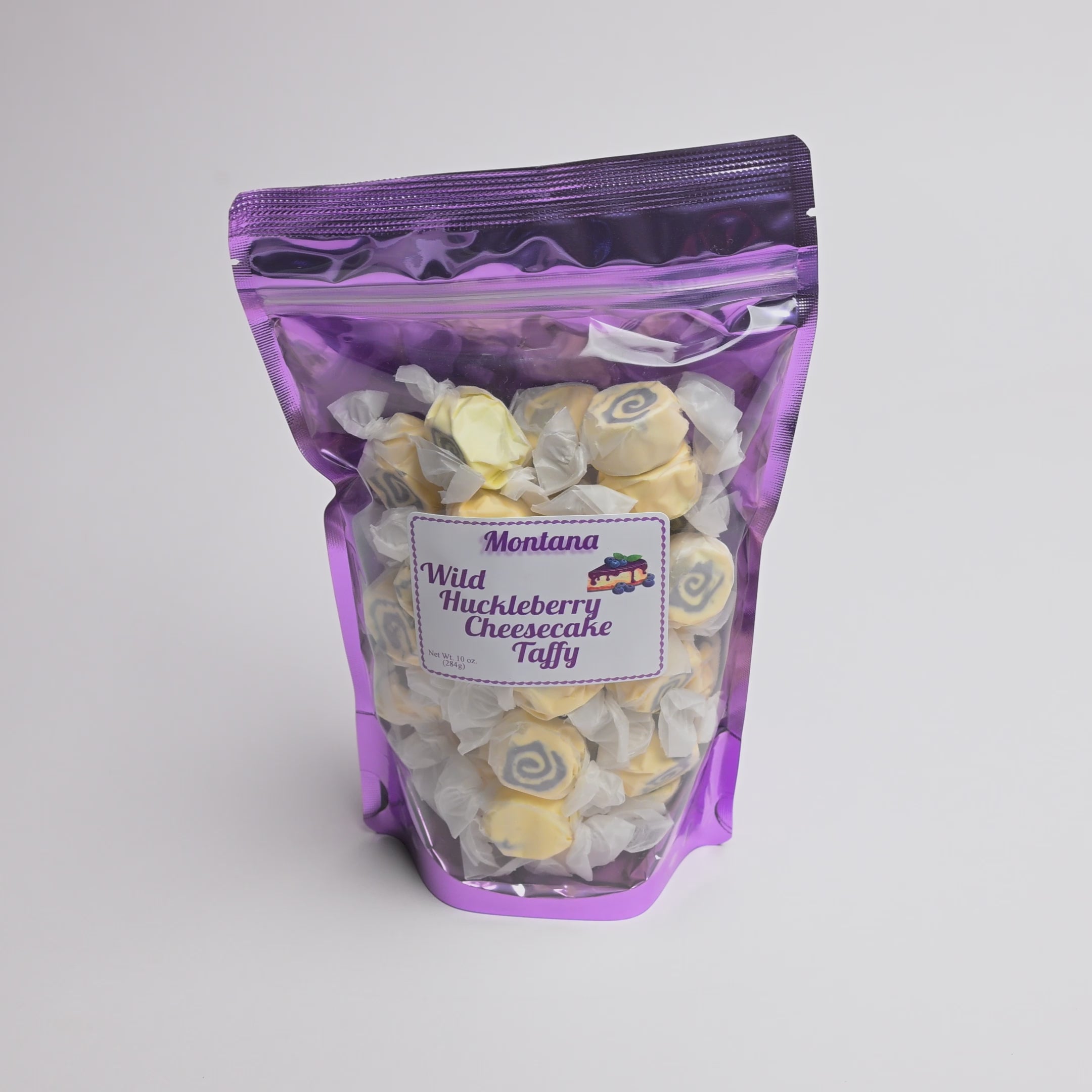 A purple resealable stand-up pouch filled with spiral-shaped Montana Wild Huckleberry Cheesecake Taffy, with a white product label on the front, photographed on a light gray background.