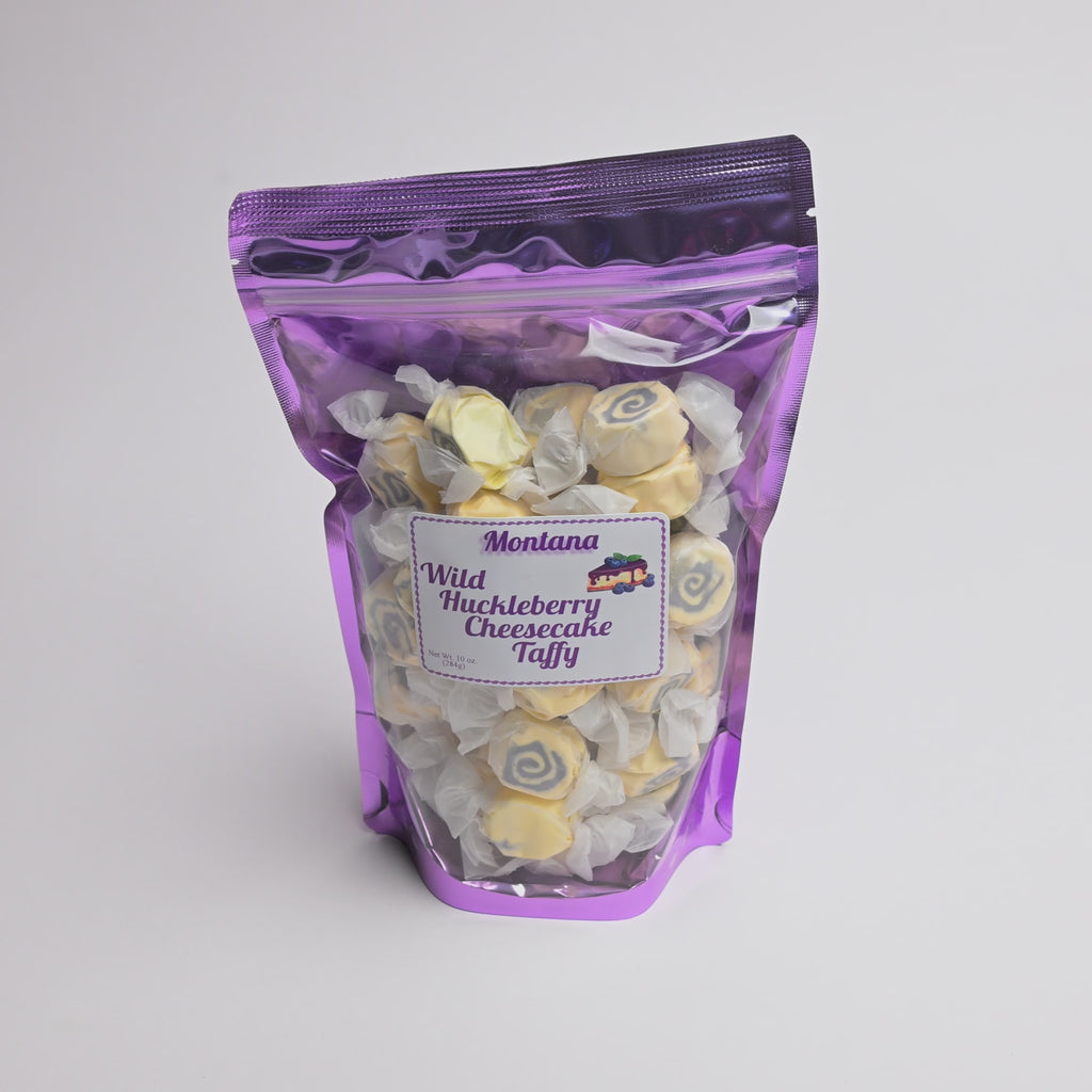 A purple resealable stand-up pouch filled with spiral-shaped Montana Wild Huckleberry Cheesecake Taffy, with a white product label on the front, photographed on a light gray background.