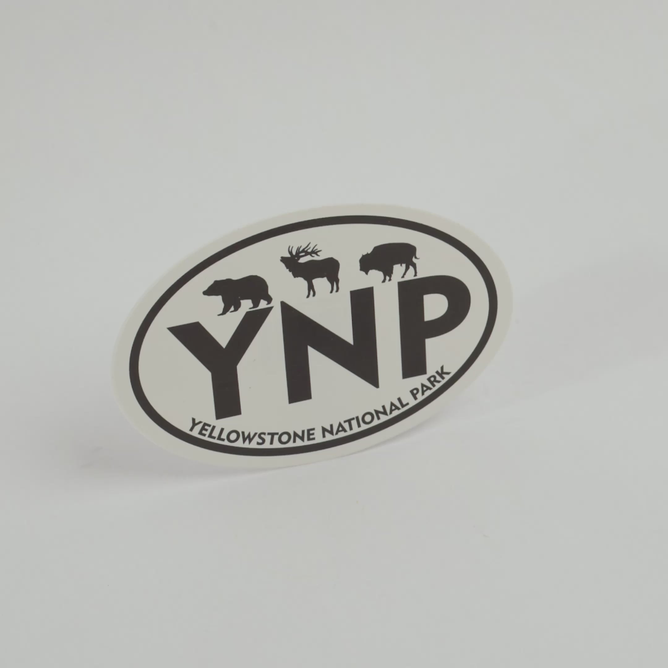 An oval Yellowstone National Park sticker featuring black silhouettes of a bear, moose, and bison above the large letters 'YNP', with 'YELLOWSTONE NATIONAL PARK' curved along the bottom border, shown on a clean white background.