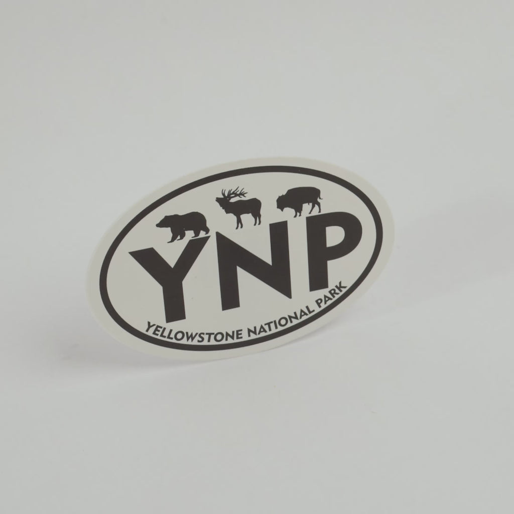 An oval Yellowstone National Park sticker featuring black silhouettes of a bear, moose, and bison above the large letters 'YNP', with 'YELLOWSTONE NATIONAL PARK' curved along the bottom border, shown on a clean white background.